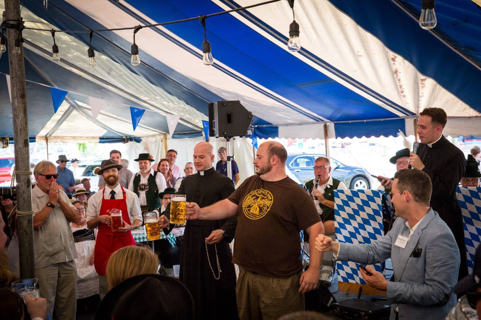 The inaugural 2017 competition under a blue-and-white striped tent: a bearded man in a brown t-shirt holds a full liter stein at arm's length beside a priest in a cassock, while a priest with a microphone speaks at right and the crowd looks on