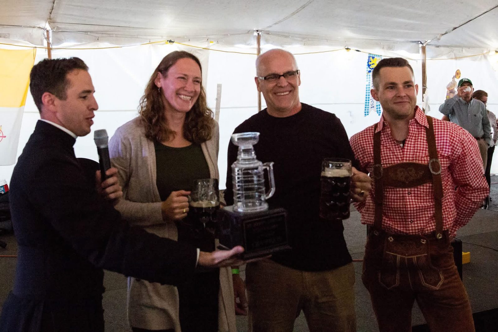 A priest with a microphone presents the Steinly Cup on stage in 2018, the year Herb Kengel and Marian Plymale won