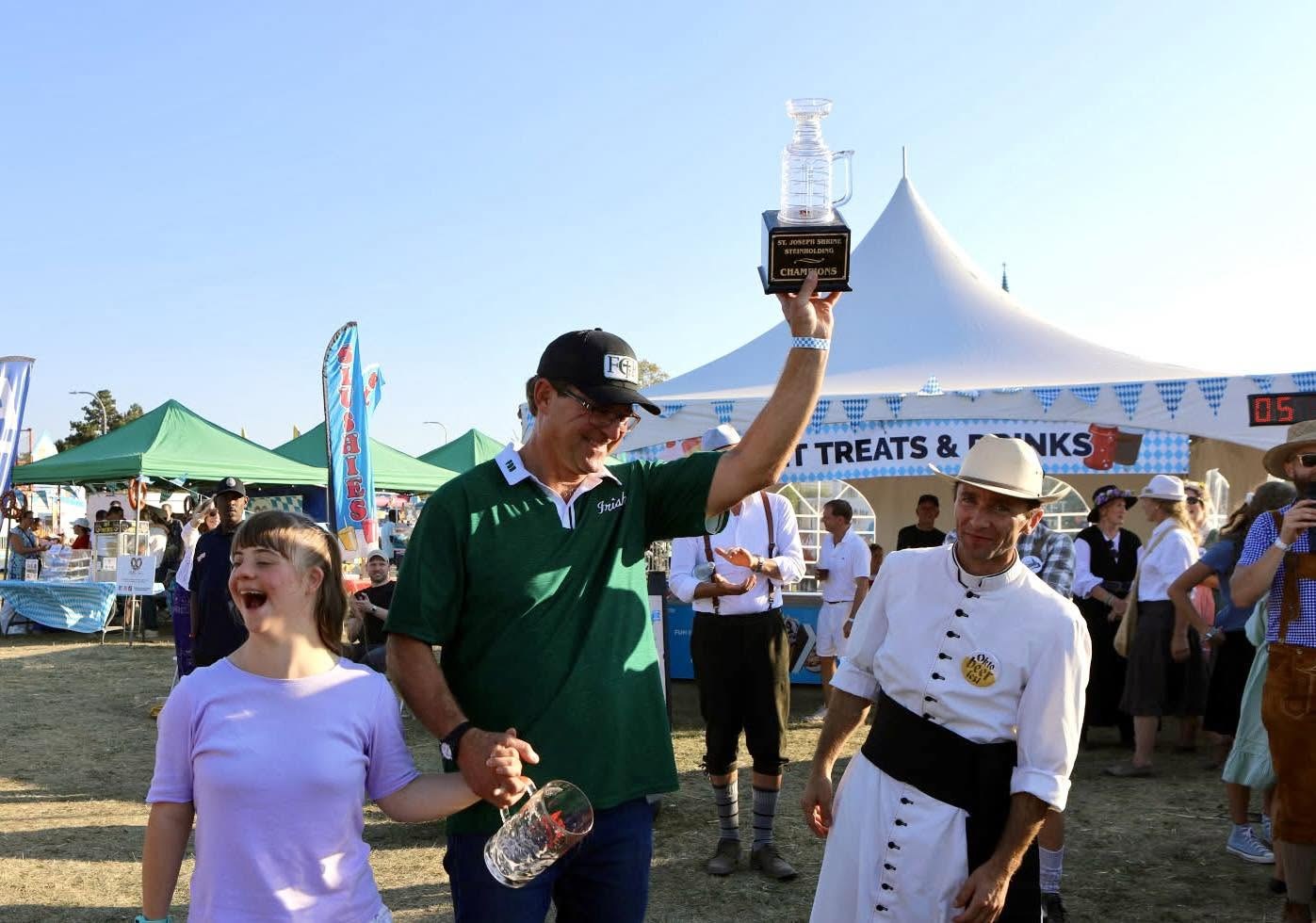2025 Steinholding champion in a green polo and cap lifting the Champions trophy beside a woman in lavender and an official in a fedora at the Sweet Treats tent