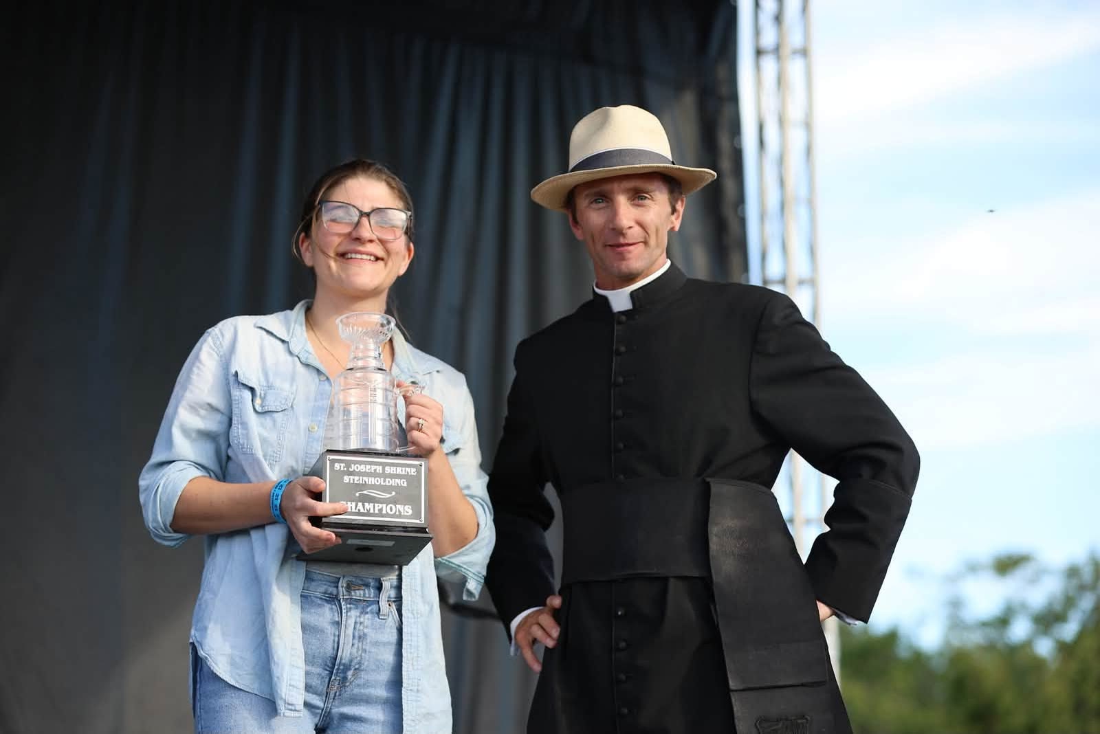 Becky Snyder, wearing glasses and denim, holding the Steinholding Champions trophy beside Canon Commins in a straw hat at DeSales Park